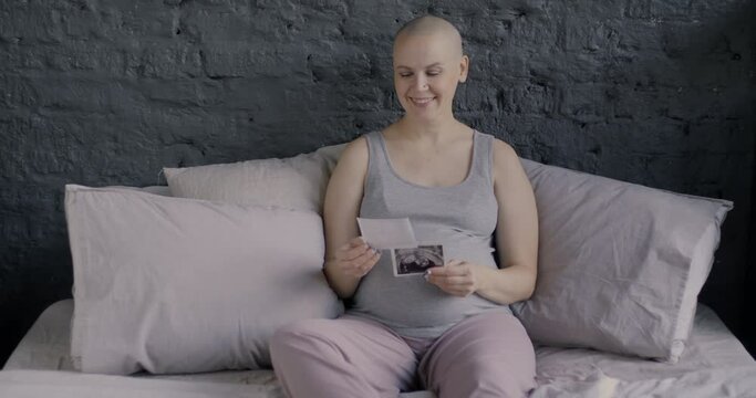 Smiling pregnant lady looking at ultrasound scans of unborn baby holding pictures dreaming about child sitting in bed at home. Healthy pregnancy and motherhood concept.