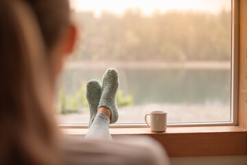 person relaxing at home in the morning feet up, warm socks,  looking out her window at the beautiful nature view feeling at peace calm - obrazy, fototapety, plakaty