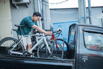 Asian man locking a bicycle while putting it on a pickup truck with a bicycle shop in the background