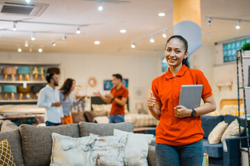 beautiful woman shows thumb holding a tablet standing against the background of buyer and shopkeeper in furniture store