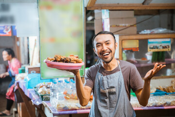 smiling Asian youth wearing an apron with one hand holding a side dish at a traditional stall