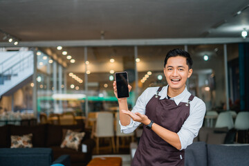 male shop assistant in apron with hand gesture presenting mobile phone in front of furniture store