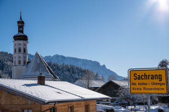 Church tower and town sign of the mountaineering village of Sachrang in Chiemgau, Bavaria, Germany with a view of the Zahmer Kaiser in Tyrol, Austria in winter