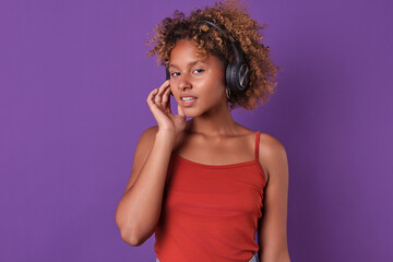 Young smiling cute African American woman teenager in wireless headphones looks at screen calling to use audio gadget to listen to educational podcasts or music stands in purple studio.