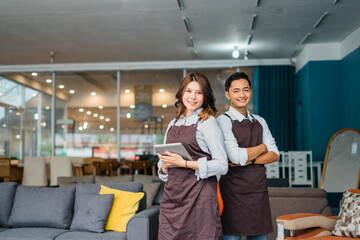 smiling male and female waitress in apron hold a tablet standing in front of couch in furniture store department