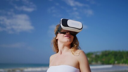 Woman immersed in VR experience on sunny beach flexes hand, explores virtual reality tropical simulation. VR headset user interacts in digital world, tactile gestures in sand, sky backdrop. Slowmo - Powered by Adobe