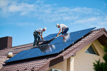 Workers building solar panel system on rooftop of house. Two men installers in helmets installing...