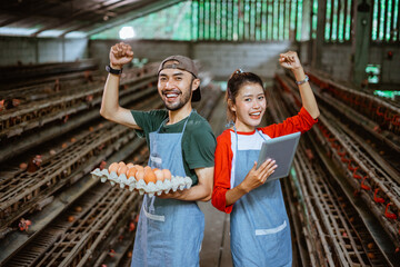 entrepreneur couple celebrate success by raising both hands with a cardboard egg tray and holding a pad standing at a poultry farm