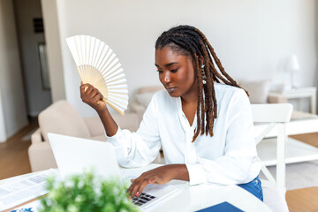 Tired African woman work on laptop at home office wave with hand fan suffer from heatstroke...