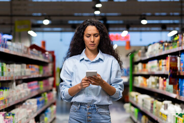 Concentrated young woman browsing on her phone while shopping in a supermarket, possibly looking up product reviews.
