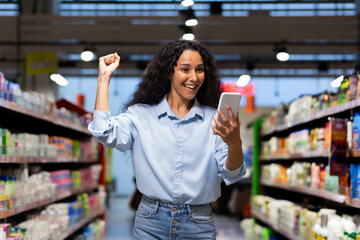 Joyful young female shopper raises her fist in victory while looking at her phone in a grocery...