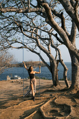 A beautiful young brunette, in a stylish dress, sits on a swing suspended on a tree, against the backdrop of the ocean.