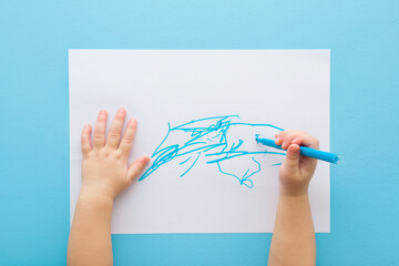 Baby boy hand holding marker and drawing first scratches lines on white paper. Blue table background. Pastel color. Closeup. Toddler development. Learning painting. Point of view shot. Top down view.