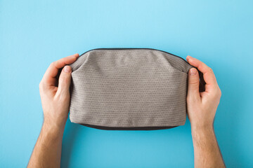 Young adult man hands holding and showing new gray cosmetic travel bag on light blue table...