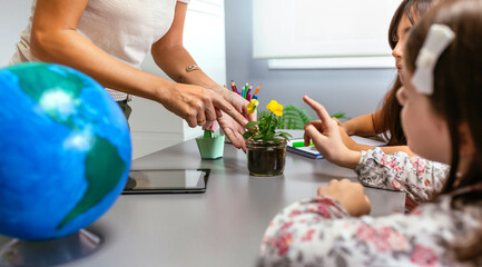Unrecognizable female teacher and young students pointing to a pansy plant inside of glass pot in ecology classroom. Botanical and natural science education concept.