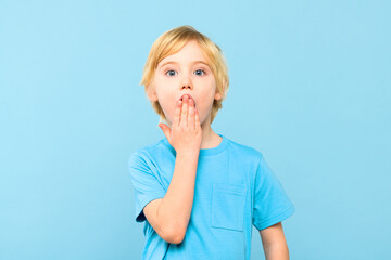 Wow! Portrait of a shocked cute little boy with blond hair covering open mouth with one hand, on...