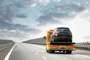 A black SUV is being transported by a yellow tow truck along an empty highway with a cloudy sky...