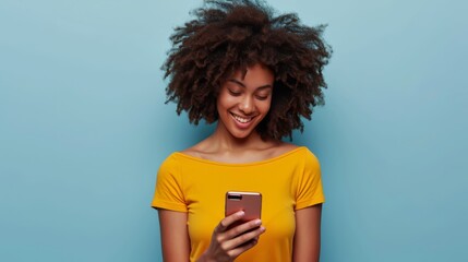 A young African American woman, radiating positivity, smiles while wearing a yellow top and using her smartphone against a light blue background - obrazy, fototapety, plakaty