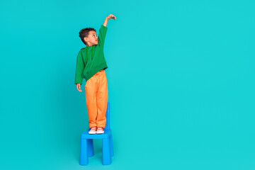 Full length photo of adorable preteen age boy standing on chair raise hand measure size product...