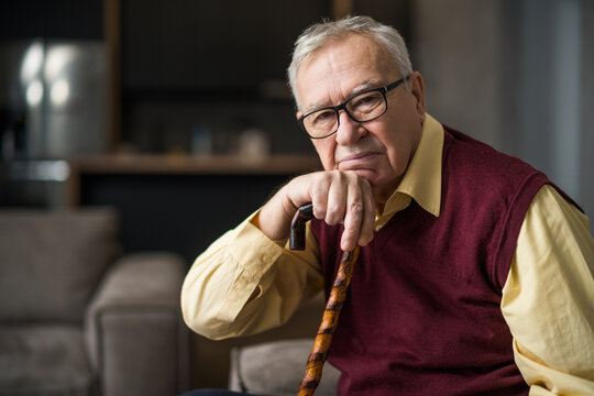 Pensive and worried senior man is sitting on bed in his home. Portrait of senior man with walking cane.