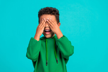 Photo of overjoyed schoolboy with wavy hair dressed green pullover arms cover eyes play peekaboo...