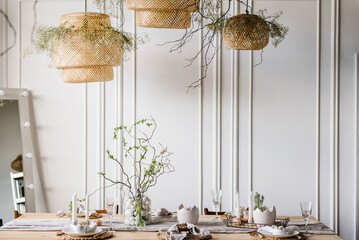 Interior design of easter dining room. Happy easter dinner with decor at home. Minimalist composition in living room with copy space, hanging lamp, candles, easter eggs, vase with twig, branch, leaves - obrazy, fototapety, plakaty