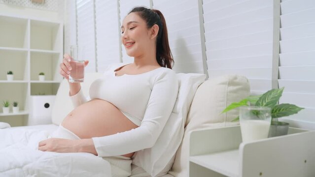 Asian pregnant woman drinking clean water between a day in bedroom at home. Asian mother sip water often and control urination while pregnancy.