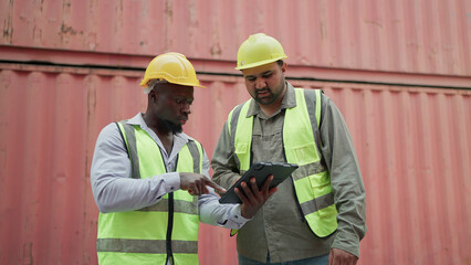 Two male container engineers working on tablet inspecting containers around a shipping yard of logistic freight forwarder company. Two engineer checking container storage with cargo container