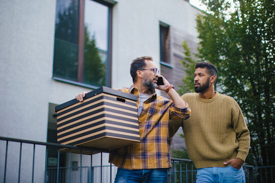 Man Helping His Friend Move Into New Apartment In Building. Man Is Moving Into New Flat, Carrying Box From The Moving Truck.