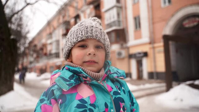 Close-up of a child girl headshot in winter clothing outdoors on city streets with snow. Knitted hat and scarf, cold weather children activities 