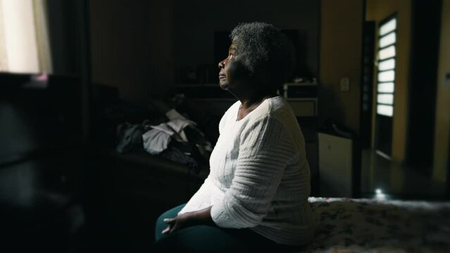 Pensive Senior African American woman in 80s seated by bedside with sad lonely look gazing at window in messy bedroom depicting solitude in old age