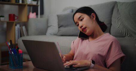 Portrait of Young woman sitting on the floor leaning against a sofa working with a laptop while talking mobile phone at home office.