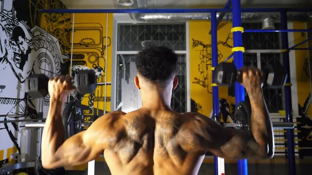 Sporty african man with tattoos lifting heavy black dumbbells at sport club. Muscular afro american sportsman pumping muscles at modern gym. Athletic black guy exercising at fitness centre. Slow mo