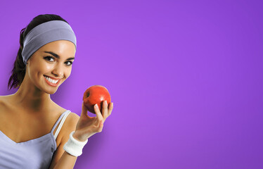 Portrait image of african american woman in sportswear with red apple, isolated on violet purple background. Young sporty model at studio. Dieting, fitness, dental care ad concept. - Powered by Adobe