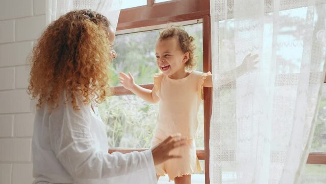 Cheerful mother playing with daughter hiding behind the curtains in living room at home. Little cute girl tease and enjoy with mother in bedroom at house. Family relationship concept.