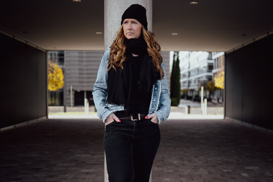 The picture shows a woman around thirty with long wavy red hair. She is dressed with black cap denim jacket and black pants scarf and sweater.