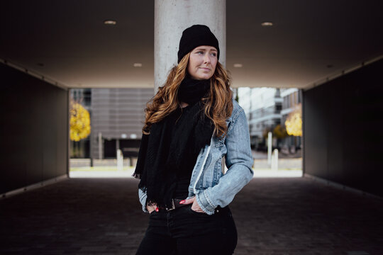 The picture shows a woman around thirty with long wavy red hair. She is dressed with black cap denim jacket and black pants scarf and sweater.