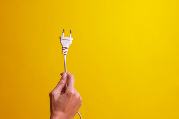 Hand holding white electrical plug isolated on yellow background with copy space. Save energy and earth hour concept - obrazy, fototapety, plakaty