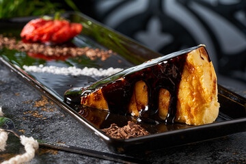 A close-up photo of a piece of cheesecake with chocolate sauce on a black plate