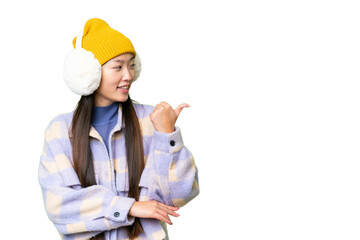 Young Asian woman wearing winter muffs over isolated chroma key background pointing to the side to present a product