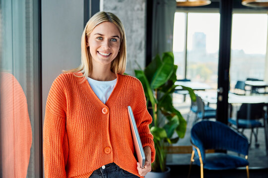 Happy young businesswoman standing with laptop near window in office