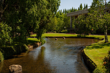 Fototapeta premium A tranquil stream meanders through a picturesque park, enveloped by a vibrant tapestry of green foliage, creating a peaceful and idyllic scene.