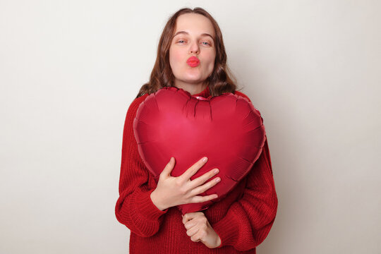 Romantic flirting brown haired adult woman wearing red sweater holding heart shape balloon sending air kissing with pout lips posing isolated over gray background