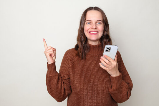 Happy smiling Caucasian woman wearing brown jumper standing isolated over gray background using mobile phone indicating away at advertisement area pointing at copy space for promotion