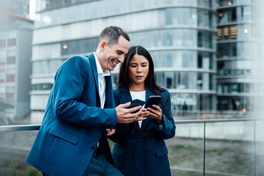Happy businessman and businesswoman using smart phone in front of buildings