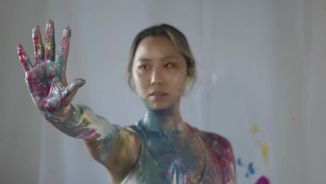 Asian woman showcases her fully painted body adorned with a spectrum of vibrant colors. Her right hand is outstretched and her palm is opened to the camera.