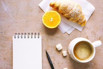 Eine Tasse Kaffee, ein frisches Croissant und ein leerer Notizblock auf einem braunen Tisch. Draufsicht.