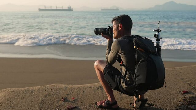 Travel photographer at the beach takes a landscape photo on his DSLR professional camera with a backpack full of tripods and other camera gear on a sunny afternoon with oil rigs in the background