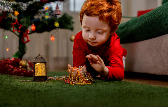 Cute redhead boy playing with Christmas ornaments on green carpet at home