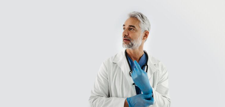 Confident Mature Doctor Putting On Medical Gloves. Handsome Doctor With Gray Hair Wearing White Coat, Stethoscope Around Neck. Banner, White Background With Copy Space.